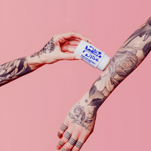Load image into Gallery viewer, Two tattooed arms against a pink background, one hand holding the full-size balm stick