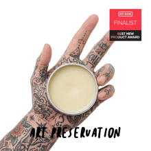 Load image into Gallery viewer, Heavily tattooed hand holding an open jar of tattoo balm, with a NY NOW Best New Product Award finalist badge
